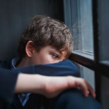 Pensive sad boy teenager in a blue shirt and jeans sitting at the window and closes his face with his hands. Les défis du refus scolaire anxieux à l’approche de la rentrée