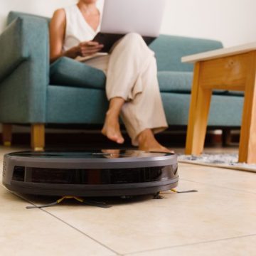 A black robot vacuum cleaner cleaning the floor and a woman with a laptop sitting on a sofa at home. La France Face au Piratage des Équipements Connectés